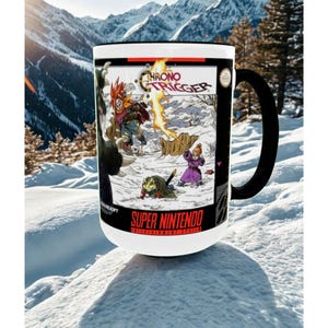 May include: White ceramic mug with a black handle featuring a colorful illustration from the video game "Chrono Trigger." The artwork depicts characters in a snowy landscape, with the words "CHRONO TRIGGER" and "SUPER NINTENDO" visible.
