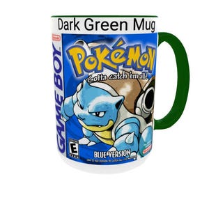 May include: A dark green mug featuring a graphic print of the Pok&eacute;mon Blue Version Game Boy game cover. The design includes the Pok&eacute;mon logo, the text Gotta catch 'em all!, the character Blastoise, and the ESRB E rating symbol on a blue background.