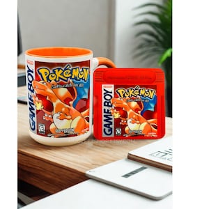 May include: A white ceramic mug with an orange interior and handle, featuring the Pok&eacute;mon Red Version Game Boy game cover art, alongside a matching red Pok&eacute;mon Red Version Game Boy game cartridge. Both items display the Charizard character.
