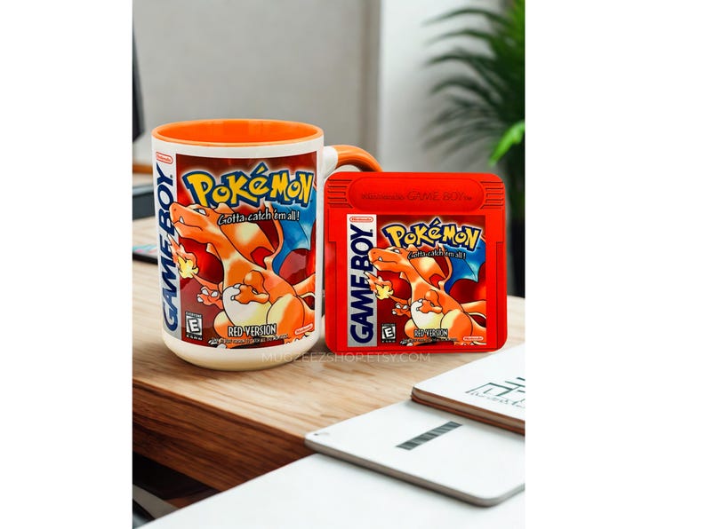May include: A white ceramic mug with an orange interior and handle, featuring the Pok&eacute;mon Red Version Game Boy game cover art, alongside a matching red Pok&eacute;mon Red Version Game Boy game cartridge. Both items display the Charizard character.