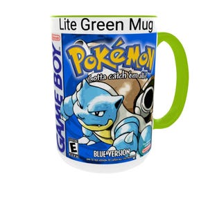 May include: A white ceramic mug with a bright lime green handle and interior rim. The exterior features a graphic print of the Pok&eacute;mon Blue Version Game Boy game box art, including the Blastoise character, the Pok&eacute;mon logo, and text.