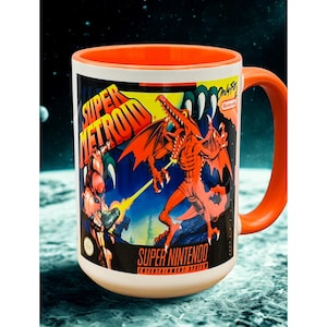 May include: A white ceramic mug with an orange interior and handle. The mug features a Super Metroid graphic with a red dragon-like creature and the text "Super Metroid" and "Super Nintendo Entertainment System". The background is a space scene.