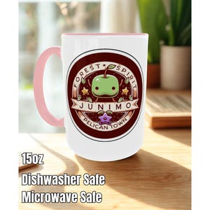 May include: A white ceramic mug with a pink handle and interior. The mug features a circular design with a green apple character and the words "Forest Spirit Junimo Pelican Town". The mug is 15oz and is dishwasher and microwave safe.