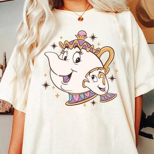 May include: Cream-colored t-shirt featuring a cartoon illustration of a teapot and teacup with smiling faces. The design includes gold, purple, and blue accents, with starburst details. The shirt is a relaxed fit.