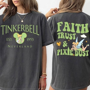 May include: Dark gray t-shirt featuring the text "TINKERBELL EST. 1953 NEVERLAND" in green. The front of the shirt has a Tinkerbell graphic inside a Mickey Mouse head. The back of the shirt has the text "FAITH TRUST & PIXIE DUST" with a Tinkerbell graphic.