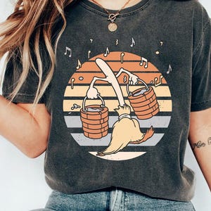 May include: Dark grey t-shirt with a cartoon broom carrying buckets, set against a sunset background. Musical notes surround the central design. A casual wear item.