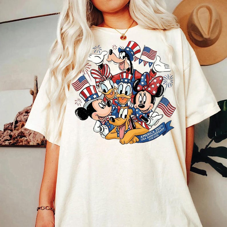 A white t-shirt with a graphic design featuring Disney characters such as Minnie Mouse, Donald Duck, and Pluto, along with American flags and a banner that reads "America 25th Anniversary".