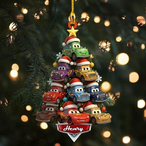 May include: A Christmas tree ornament featuring a stack of cartoon cars, each wearing a Santa hat. The cars are various colors, including green, purple, red, and blue. The ornament has a gold star at the top and a banner that says "Henry".