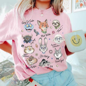 A person wearing a pink t-shirt with various cartoon animal faces and names printed on it, holding a coffee mug with a smiley face.