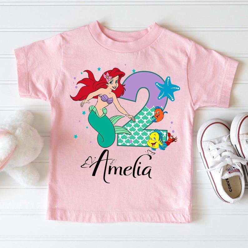 A pink t-shirt with a cartoon image of a mermaid character and the name "Amelia" printed on it, along with a starfish and other sea creatures.