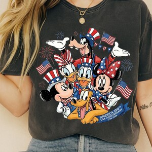 A t-shirt with a graphic design featuring Disney characters such as Minnie Mouse, Donald Duck, Goofy, and Pluto, along with American flags and fireworks.