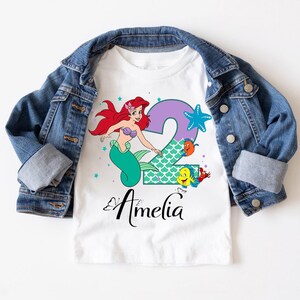 A white t-shirt with a cartoon image of a mermaid character and the number 2, along with the text "Amelia" printed on it.