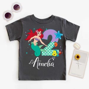 A gray t-shirt with a design featuring a mermaid character and the number 2, along with other sea creatures. The t-shirt is placed on a white surface, accompanied by a pair of sunglasses, a small flower, and a card.
