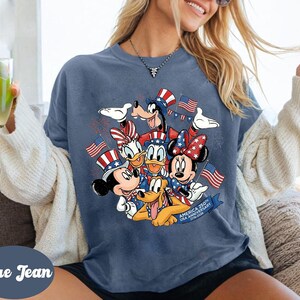 A woman wearing a blue t-shirt with a graphic of Disney characters, including Goofy, Minnie Mouse, Donald Duck, and Pluto, along with American flags. She is holding a glass of lemonade and appears to be enjoying the moment.