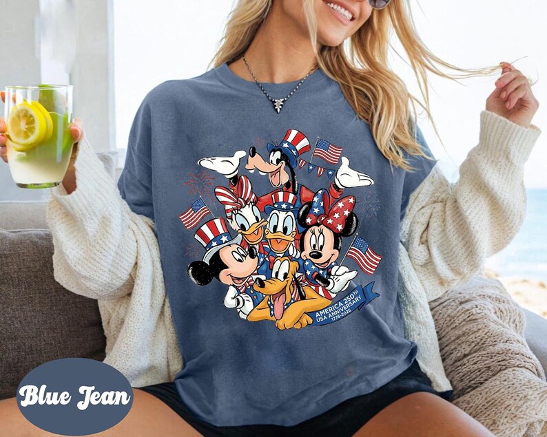 A woman wearing a blue t-shirt with a graphic of Disney characters, including Goofy, Minnie Mouse, Donald Duck, and Pluto, along with American flags. She is holding a glass of lemonade and appears to be enjoying the moment.