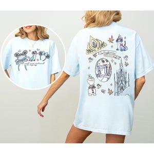 Peut inclure: T-shirt bleu clair avec des graphiques sur le thème de Disney. L'avant affiche des oreilles de Mickey Mouse, une grenouille et des oreilles de Minnie Mouse avec le texte "The cold never bothered me anyway." Le dos a une couronne, un château et d'autres illustrations.