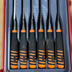 May include: A set of six chisels in a red and grey case. The chisels are labelled with their size in millimetres and inches. The case has the text "A+ DESIGNS" printed on it.