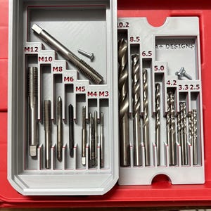 May include: A grey plastic organiser with two rows of tools. The top row contains eight tap and die sets labelled M12, M10, M8, M6, M5, M4, and M3. The bottom row contains ten drill bits labelled 10.2, 8.5, 6.5, 5.0, 4.2, 3.3, and 2.5. The organiser is labelled "A+ DESIGNS" on both sides.