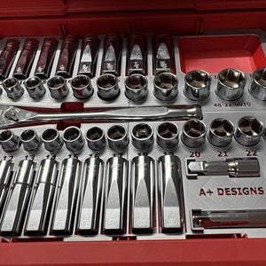 May include: A red toolbox drawer containing a set of chrome sockets, ratchets, and extensions. The tools are neatly arranged in a grey tray. The ratchet is branded "Milwaukee". The tray has the text "A+ DESIGNS".