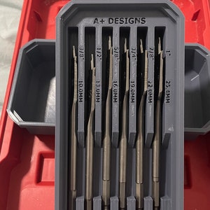 May include: A grey plastic storage case with the text "A+ DESIGNS" on the front. The case holds a set of seven drill bits, each with a different size marked in millimetres: 10.0mm, 13.0mm, 16.0mm, 19.0mm, 22.0mm, and 25.4mm.