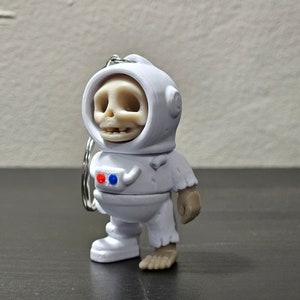 May include: White astronaut figurine with a skeleton head and a red and blue detail on the chest. The figurine is wearing a white spacesuit with a helmet and has a silver keychain attached.