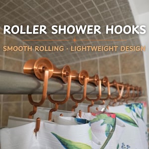 Op de afbeelding: Koperkleurige douchegordijnrollers op een douchestang, met een wit douchegordijn met bloemenpatroon. De tekst "ROLLER SHOWER HOOKS" staat boven de rollers, met "SMOOTH ROLLING • LIGHTWEIGHT DESIGN" eronder.