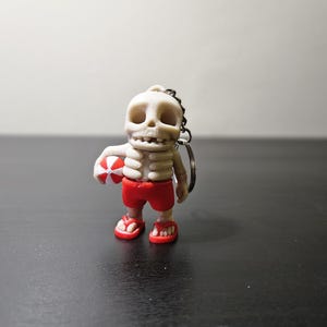 Peut inclure: Une figurine de squelette blanche portant un short de bain rouge et tenant un ballon de plage. La figurine est attachée à un porte-clés argenté.