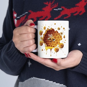 May include: White ceramic mug with a graphic of a honeybee flying out of a honeycomb with dripping honey. The honeycomb is surrounded by white flowers.