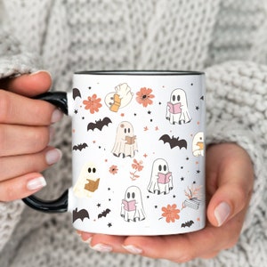 Op de afbeelding: Witte keramische mok met een zwart handvat en rand, met een herhalend patroon van stripgeesten die boeken lezen, vleermuizen, bloemen en sterren in tinten perzik, bruin en zwart. De mok wordt vastgehouden door een persoon die een witte trui draagt.