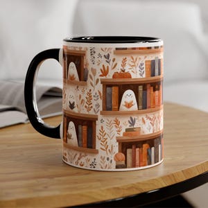 Może przedstawiać: Ceramiczny kubek z czarnym uchwytem i obramowaniem. Kubek ma powtarzający się wzór półek z książkami z duchami czytającymi książki. Tło ma jesienny motyw z liśćmi i dyniami.