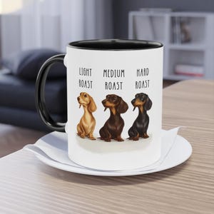 May include: A white ceramic coffee mug with a black handle and rim, featuring illustrations of three dachshunds representing light, medium, and hard coffee roasts. The mug sits on a white saucer.
