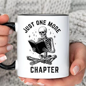 May include: White ceramic mug with a black handle and rim. The mug features a black and white illustration of a skeleton reading a book, with the text "JUST ONE MORE CHAPTER" in a gothic font. The mug is held by a person.