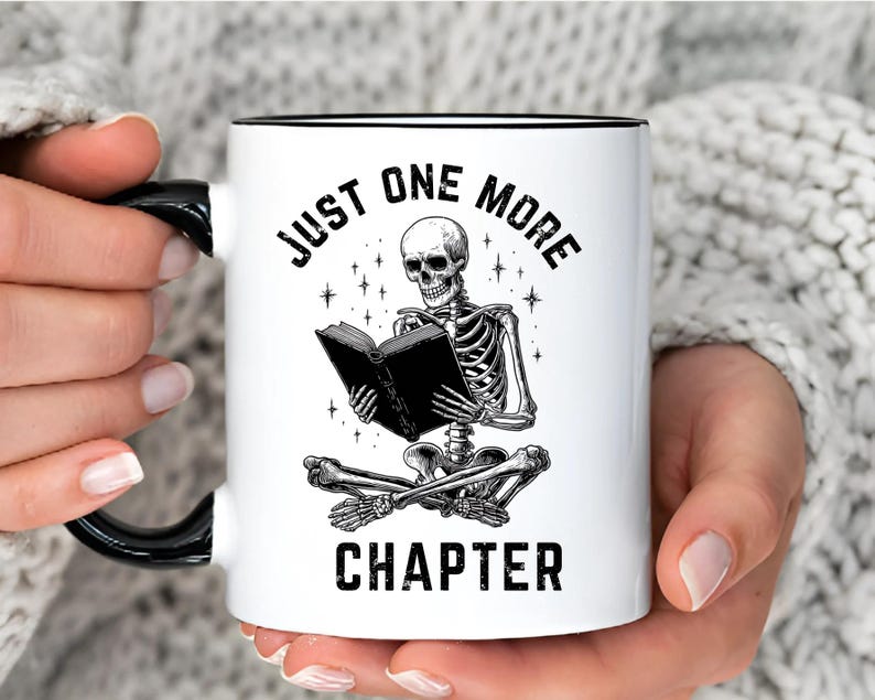 May include: White ceramic mug with a black handle and rim. The mug features a black and white illustration of a skeleton reading a book, with the text "JUST ONE MORE CHAPTER" in a gothic font. The mug is held by a person.