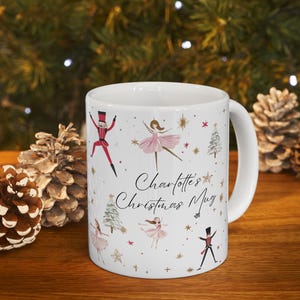 May include: White ceramic mug with a handle, featuring a Christmas theme. The mug is decorated with illustrations of ballerinas in pink tutus, toy soldiers, Christmas trees, and stars. The text "Charlotte's Christmas Mug" is also printed on the mug.