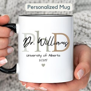 Peut inclure: Mug en céramique blanche avec une anse et un bord noirs, portant l'inscription "Personalized Mug". Le mug affiche "PhD Dr. Williams" dans une police élégante, ainsi que "University of Alberta 2025" et un petit symbole de cœur.