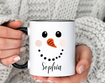May include: White ceramic mug featuring a snowman face design with black eyes, an orange carrot nose, and pink cheeks. The mug has a black handle and rim, with the name "Sophia" in a script font below the snowman's smile.