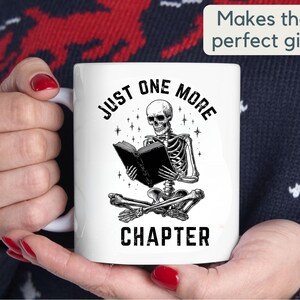 May include: White ceramic mug featuring a black and white illustration of a skeleton reading a book. The text on the mug reads "JUST ONE MORE CHAPTER". The mug also has the text "Makes the perfect gift".