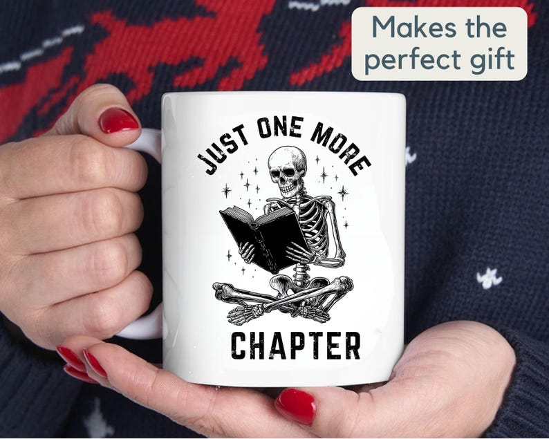 May include: White ceramic mug featuring a black and white illustration of a skeleton reading a book. The text on the mug reads "JUST ONE MORE CHAPTER". The mug also has the text "Makes the perfect gift".