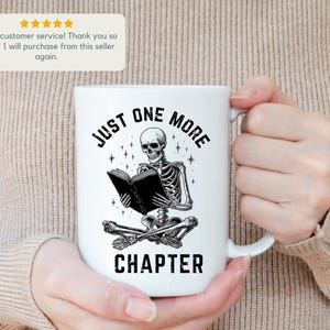 May include: White ceramic mug featuring a black and white illustration of a skeleton reading a book. The text on the mug reads "JUST ONE MORE CHAPTER". The mug has a curved handle and is held by a person.