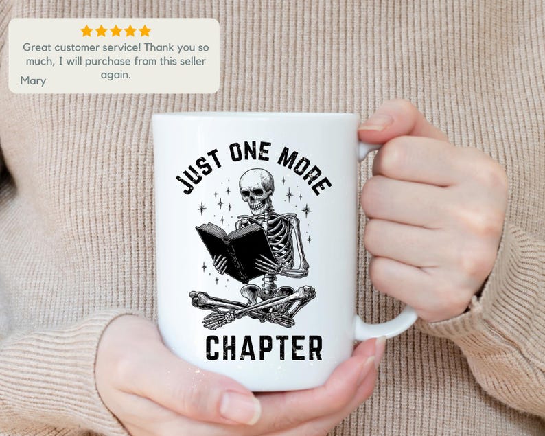 May include: White ceramic mug featuring a black and white illustration of a skeleton reading a book. The text on the mug reads "JUST ONE MORE CHAPTER". The mug has a curved handle and is held by a person.