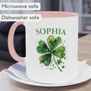 K&ouml;nnte beinhalten: Wei&szlig;e Keramiktasse mit rosafarbenem Rand, verziert mit einem gr&uuml;nen vierbl&auml;ttrigen Kleeblatt und dem Namen "SOPHIA" in schwarzer Schrift. Die Tasse ist mikrowellen- und sp&uuml;lmaschinenfest.