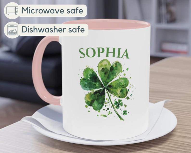 K&ouml;nnte beinhalten: Wei&szlig;e Keramiktasse mit rosafarbenem Rand, verziert mit einem gr&uuml;nen vierbl&auml;ttrigen Kleeblatt und dem Namen "SOPHIA" in schwarzer Schrift. Die Tasse ist mikrowellen- und sp&uuml;lmaschinenfest.