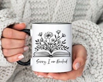 May include: White ceramic mug with a black rim and handle. The mug features a black and white illustration of an open book with flowers growing from it. The text "Sorry, I am Booked" is printed below the book.