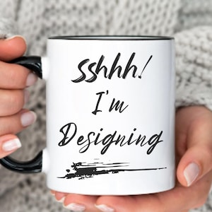 Könnte beinhalten: Weiße Keramiktasse mit schwarzem Rand und Henkel. Die Tasse hat einen schwarzen Text mit der Aufschrift "Sshhh! I'm Designing" mit einem schwarzen Pinselstrich darunter.