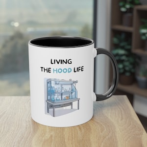 May include: White ceramic mug with a black rim and handle. The mug features the text "Living the Hood Life" and a cartoon illustration of a laboratory fume hood with glassware.