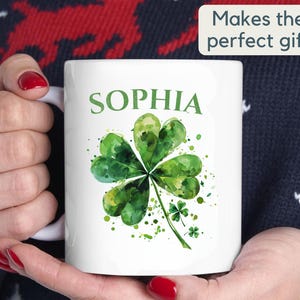 K&ouml;nnte beinhalten: Wei&szlig;e Keramiktasse mit einem gr&uuml;nen Aquarell-Kleeblatt-Design und dem Namen "SOPHIA" in Schwarz gedruckt.