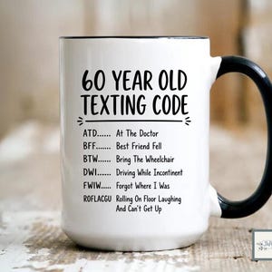 May include: White ceramic mug with a black handle and rim. The mug features the text "60 YEAR OLD TEXTING CODE" with a list of acronyms and their meanings, such as "ATD... At The Doctor".