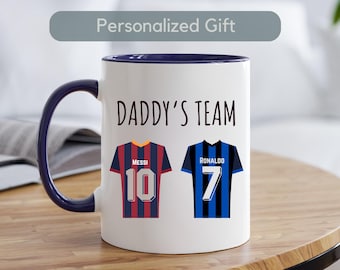 Op de afbeelding: Een witte keramische mok met een blauwe rand. De mok heeft een afbeelding van twee voetbalshirts met de tekst "Daddy's Team" boven de shirts. Het shirt aan de linkerkant heeft de naam "Messi" en het nummer "10". Het shirt aan de rechterkant heeft de naam "Ronaldo" en het nummer "7".