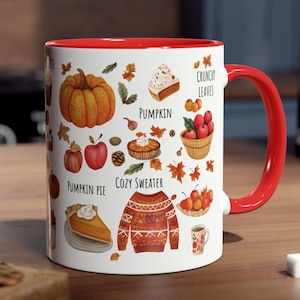 Op de afbeelding: Witte keramische mok met een rode binnenkant en handvat, versierd met herfstthema-illustraties. Het ontwerp bevat een pompoen, appel, pompoentaart en de woorden "Crunchy Leaves" en "Cozy Sweater."