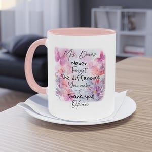 May include: White ceramic mug with a pink interior and handle, resting on a white saucer. The mug features a floral design with butterflies and the text "Mrs. Davis, Never Forget The difference You make, Thank you! Olivia."